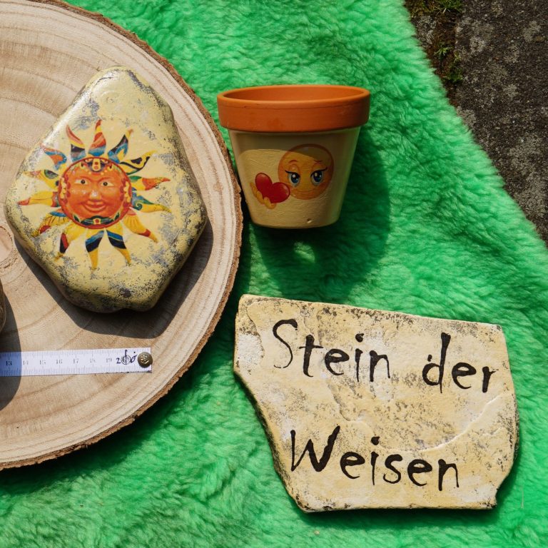 Bunte Steine und ein Blumentopf mit einer Zeichnung auf grünem Untergrund.